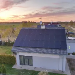 Dom jednorodzinny z panelami słonecznymi na dachu, widok z góry, w tle zachodzące słońce i krajobraz wiejski.