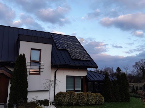 Dom jednorodzinny z panelami słonecznymi na dachu, widok z ogrodu, niebo z chmurami w tle.