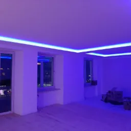 Wnętrze pokoju z białymi ścianami i podłogą z jasnego drewna, oświetlone niebieskim światłem LED zamontowanym pod sufitem, widoczne okna z widokiem na miasto nocą oraz kartony na podłodze.