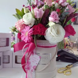 Kompozycja kwiatowa w okrągłym pudełku z różowymi wstążkami i bilecikiem, otoczona pudełkami świec zapachowych, obok leżą złote nożyczki.