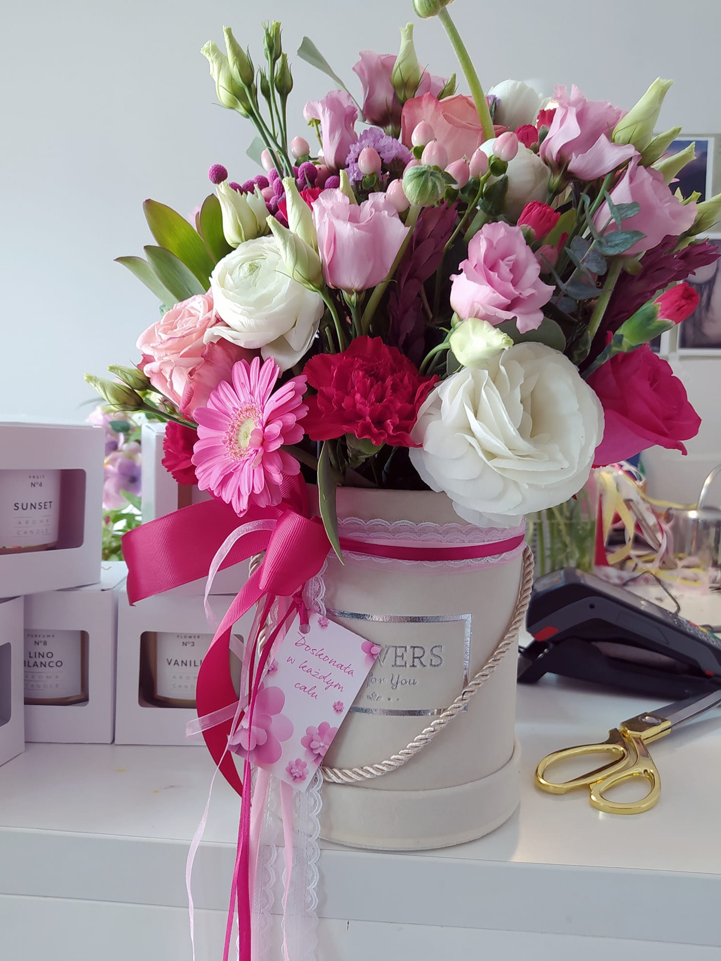 Kompozycja kwiatowa w okrągłym pudełku z różowymi wstążkami i bilecikiem, otoczona pudełkami świec zapachowych, obok leżą złote nożyczki.
