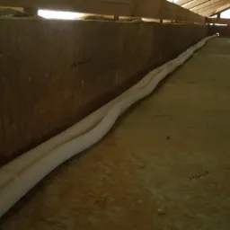 Dwie izolowane rury prowadzone wzdłuż ściany w surowym pomieszczeniu poddasza z drewnianą konstrukcją dachu i oknem dachowym w tle.