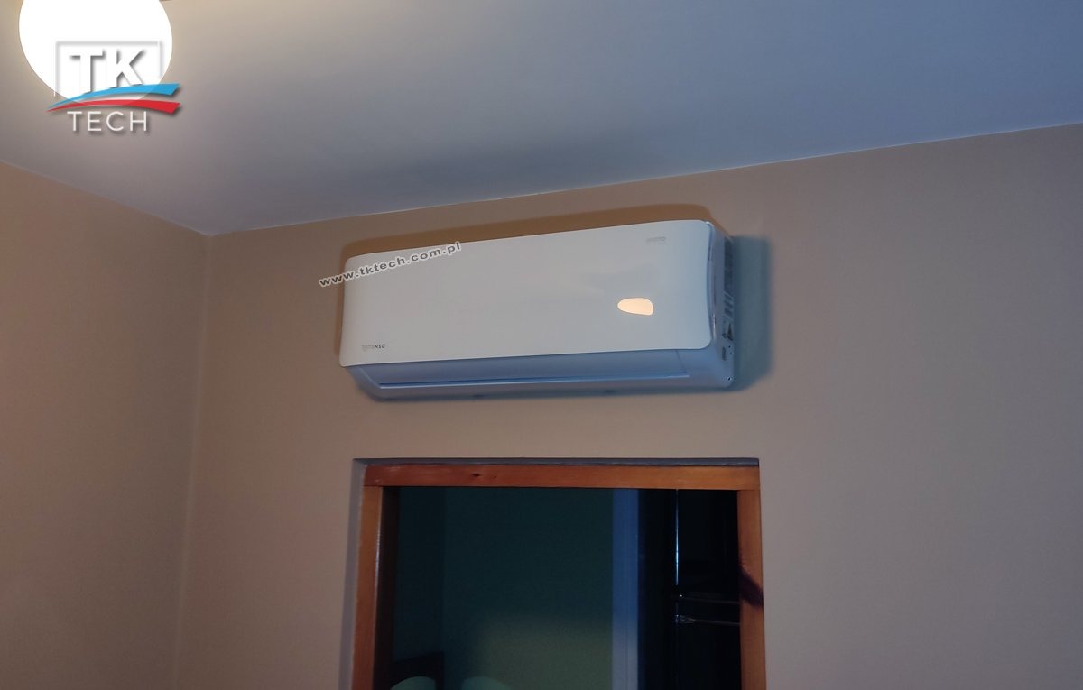 Biały klimatyzator ścienny zamontowany nad drewnianą futryną drzwiową w pokoju o beżowych ścianach, widoczne logo firmy TK Tech w lewym górnym rogu.