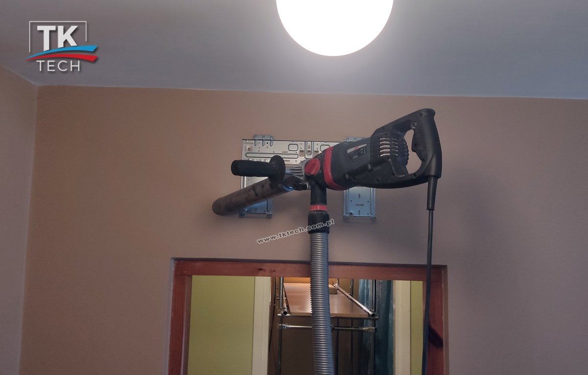 Wiercenie otworu w ścianie pod instalację klimatyzacji za pomocą wiertnicy z odsysaniem pyłu, widoczne uchwyty montażowe na ścianie i fragment ramy drzwiowej.