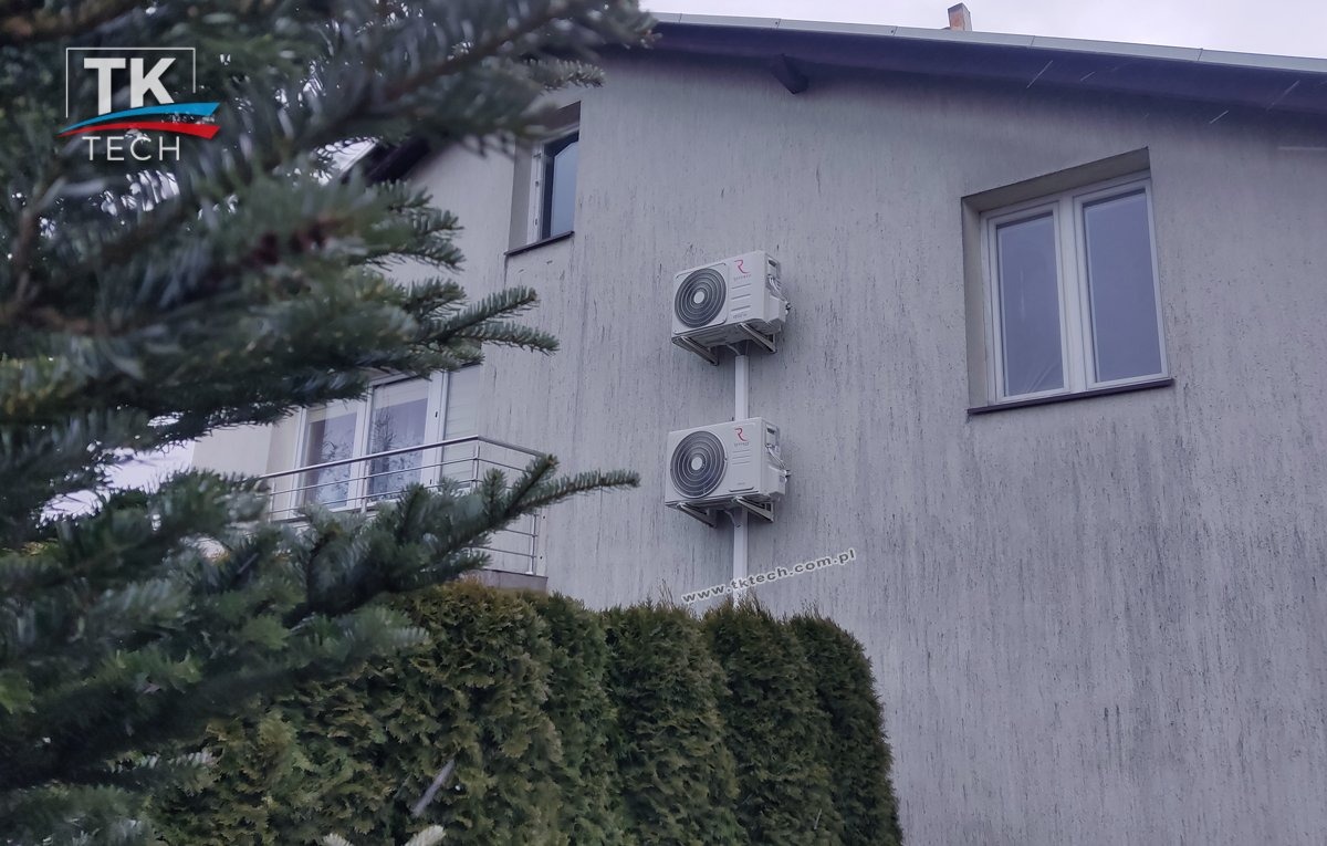 Dwa klimatyzatory zamontowane jeden nad drugim na elewacji budynku, widoczne zza gęstego żywopłotu i gałęzi drzewa iglastego. Na ścianie widoczne zacieki i okno.