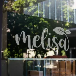 Biały napis 'Melisa' z motywem liścia na szklanej witrynie sklepu, odbijający zieleń drzew, z widocznymi produktami w tle.