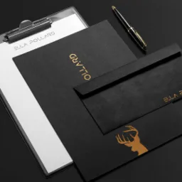 Czarny papier firmowy z logo i złotym tekstem na desce z klipsem, koperta i długopis na ciemnym tle.