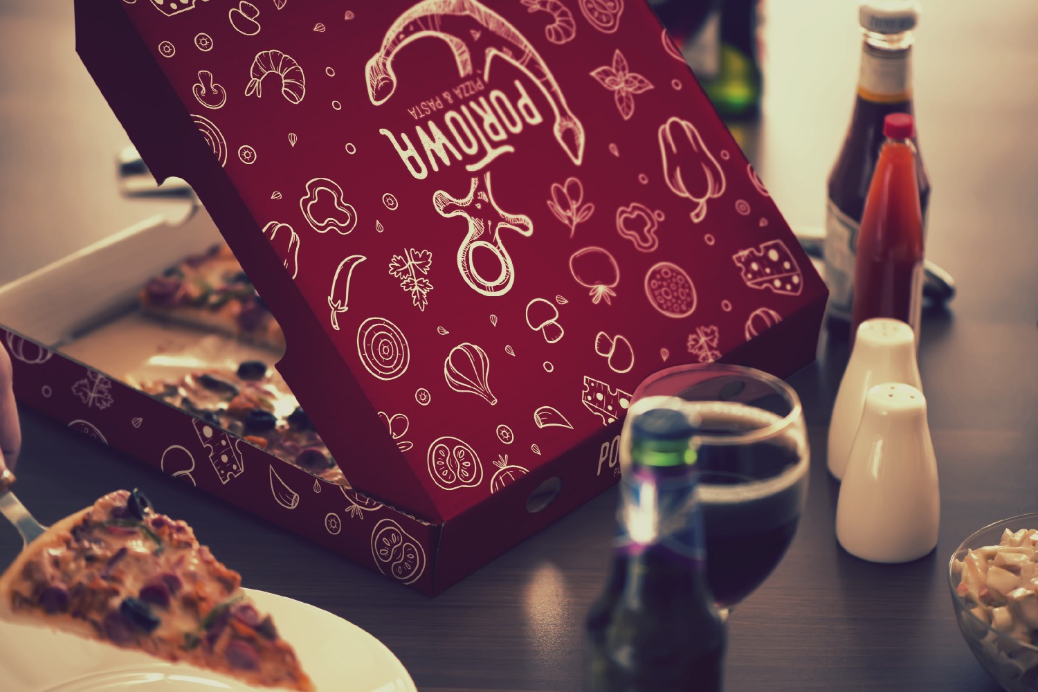 Otwarta, bordowa pizza w pudełku z białymi rysunkami warzyw i sera, z logotypem firmy na wieczku, obok szklanka z napojem, butelki z sosem i miseczka sałatki na drewnianym stole, podczas nakładania...