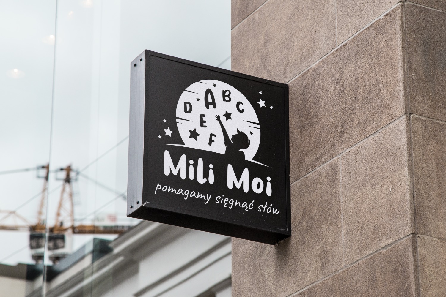 Czarny, kwadratowy szyld reklamowy na ścianie budynku, z białym logo przedstawiającym sylwetkę dziecka sięgającego do liter w okręgu, otoczonego gwiazdami, z napisem 'Mili Moi pomagamy sięgnąć...