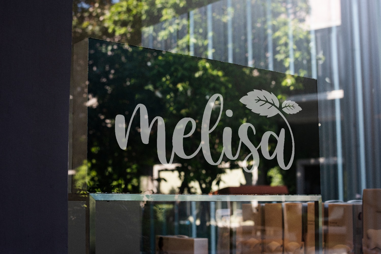 Biały napis 'Melisa' z motywem liścia na szklanej witrynie sklepu, odbijający zieleń drzew, z widocznymi produktami w tle.