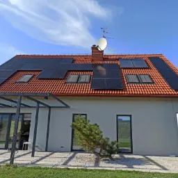 Dom jednorodzinny z panelami fotowoltaicznymi na czerwonej dachówce, widoczna antena satelitarna, błękitne niebo z delikatnymi chmurami oraz zadbany trawnik i ozdobne drzewko iglaste.