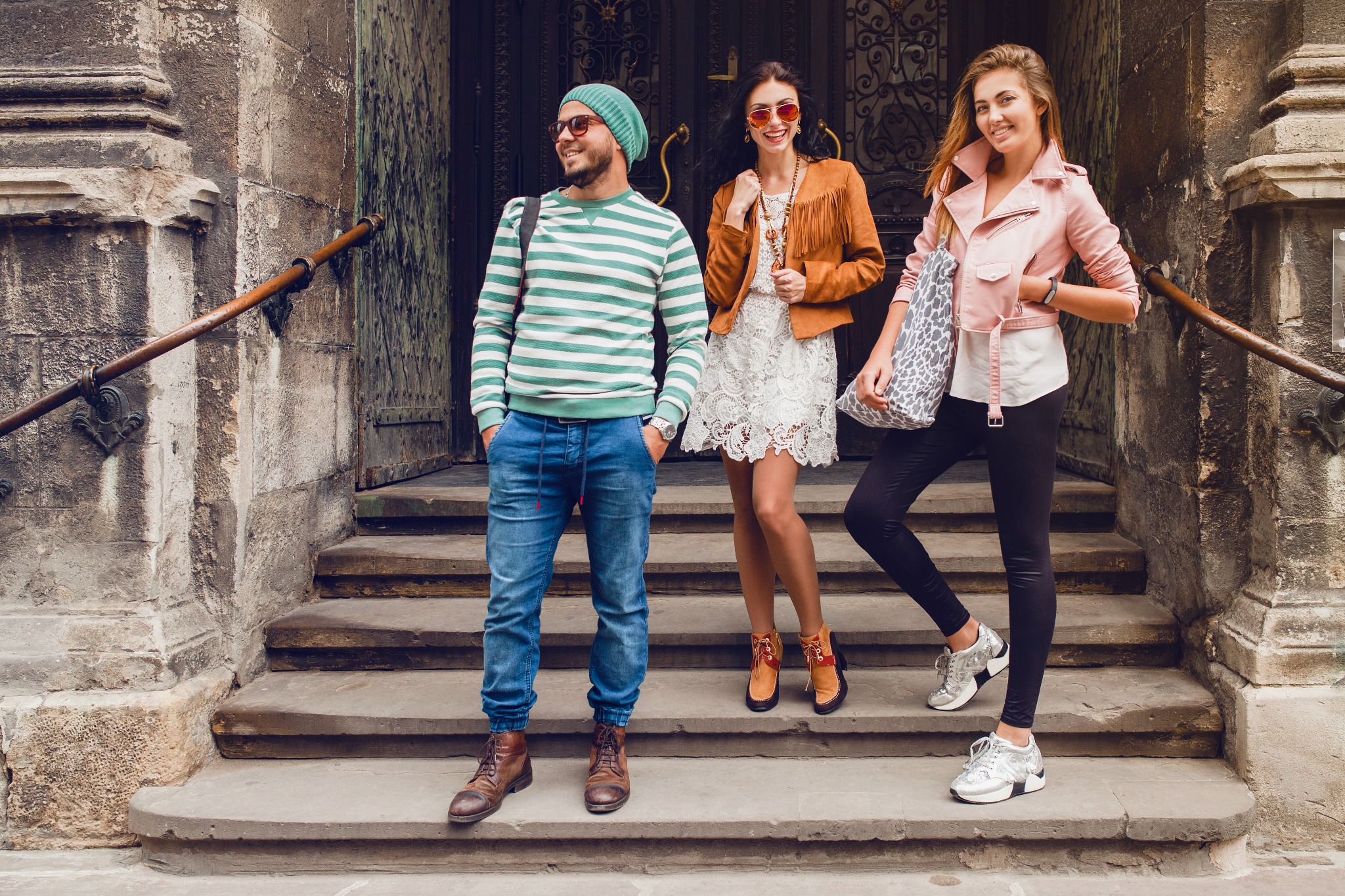 Trzy osoby ubrane w stylu miejskim pozują na kamiennych schodach przed ozdobnymi, ciemnymi drzwiami.