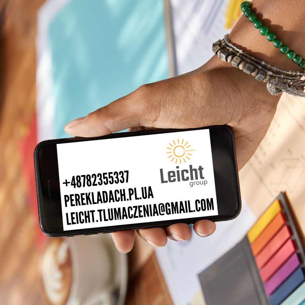 Dłoń trzymająca smartfon z wyświetlonymi danymi kontaktowymi firmy Leicht Group: numer telefonu, adres strony internetowej i adres e-mail. W tle rozmyte biurko z kolorowymi karteczkami i notatkami.