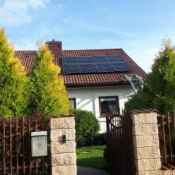 Dom jednorodzinny z panelami fotowoltaicznymi na dachu, drabina oparta o dach, widok zza ogrodzenia z tujami.