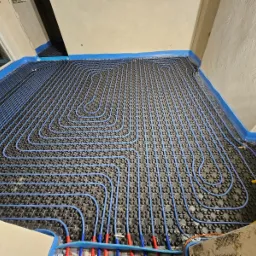 Niebieskie rury ogrzewania podłogowego ułożone spiralnie na czarnej macie izolacyjnej, zabezpieczone niebieską folią przy ścianach w surowym wnętrzu.