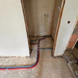SANITKAN Sylwester Iwanicki - Rury instalacji wodnej (czerwona i niebieska) ułożone na surowym podłożu w pomieszczeniu w trakcie remontu. Widoczne ślady po kuciu w ścianie.