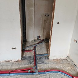 SANITKAN Sylwester Iwanicki - Instalacja rur w kolorze niebieskim i czerwonym na podłodze w surowym stanie. Widoczne otwory w ścianach i pionowa rura. Prace hydrauliczne w trakcie realizacji.