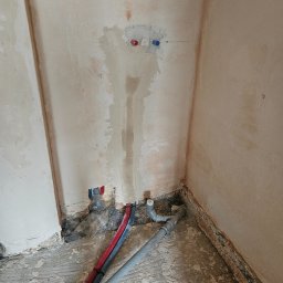 SANITKAN Sylwester Iwanicki - Fragment ściany z instalacją wodną w trakcie montażu. Widoczne rury, przyłącza i ślady po kuciu w tynku. Surowy stan wykończenia.