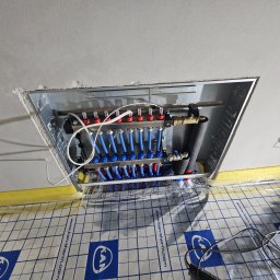 SANITKAN Sylwester Iwanicki - Rozdzielacz ogrzewania podłogowego w metalowej obudowie, widoczne niebieskie rurki i czerwone zawory, ułożony na folii izolacyjnej z nadrukiem.