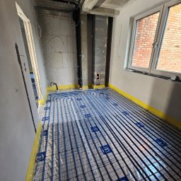 SANITKAN Sylwester Iwanicki - Niebieskie rury ogrzewania podłogowego ułożone na folii z nadrukiem w surowym wnętrzu z oknem i widoczną instalacją pod sufitem. Żółta taśma na ścianach.