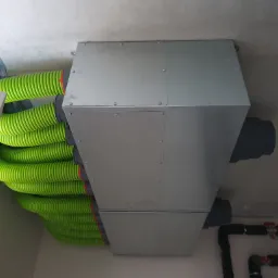 Szara skrzynka wentylacyjna z podłączonymi zielonymi, elastycznymi przewodami wentylacyjnymi, zamocowana do sufitu, z widocznymi elementami instalacji wodnej w tle.