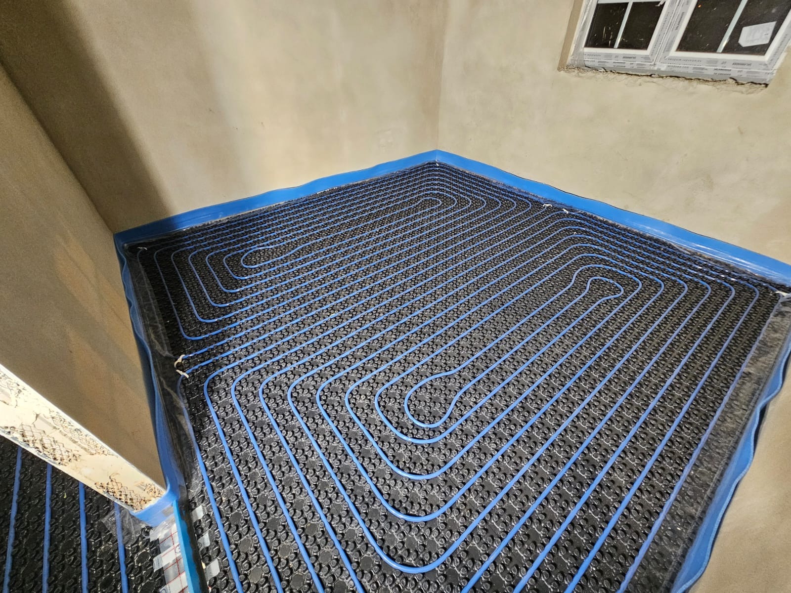 Niebieskie rury ogrzewania podłogowego ułożone na czarnej macie izolacyjnej w rogu pokoju ze ścianami w stanie surowym i oknem w tle.