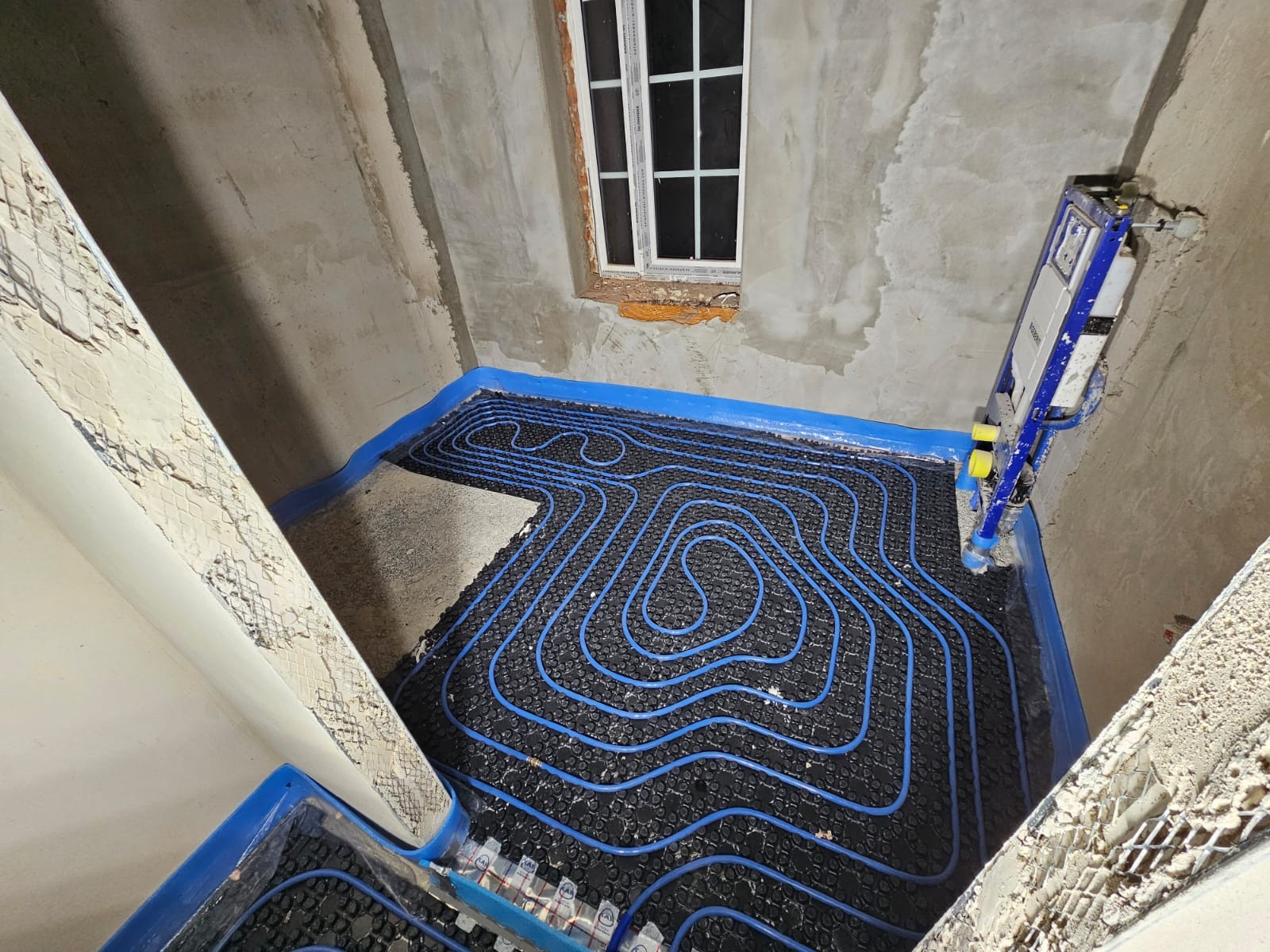 Niedokończona instalacja ogrzewania podłogowego z niebieskimi rurkami ułożonymi na czarnej macie w surowym pomieszczeniu z oknem i widocznym stelażem podtynkowym WC.