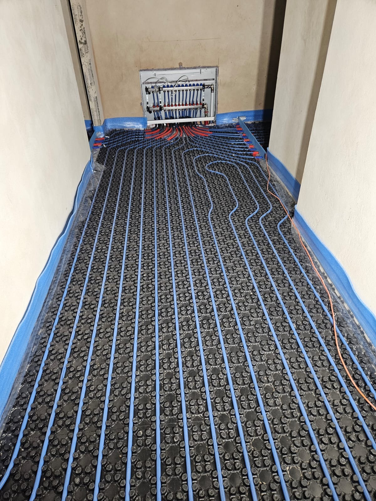 Niedokończona instalacja ogrzewania podłogowego z niebieskimi rurami na czarnej macie izolacyjnej, widoczna rozdzielnia z przewodami w tle, zabezpieczone ściany niebieską folią.