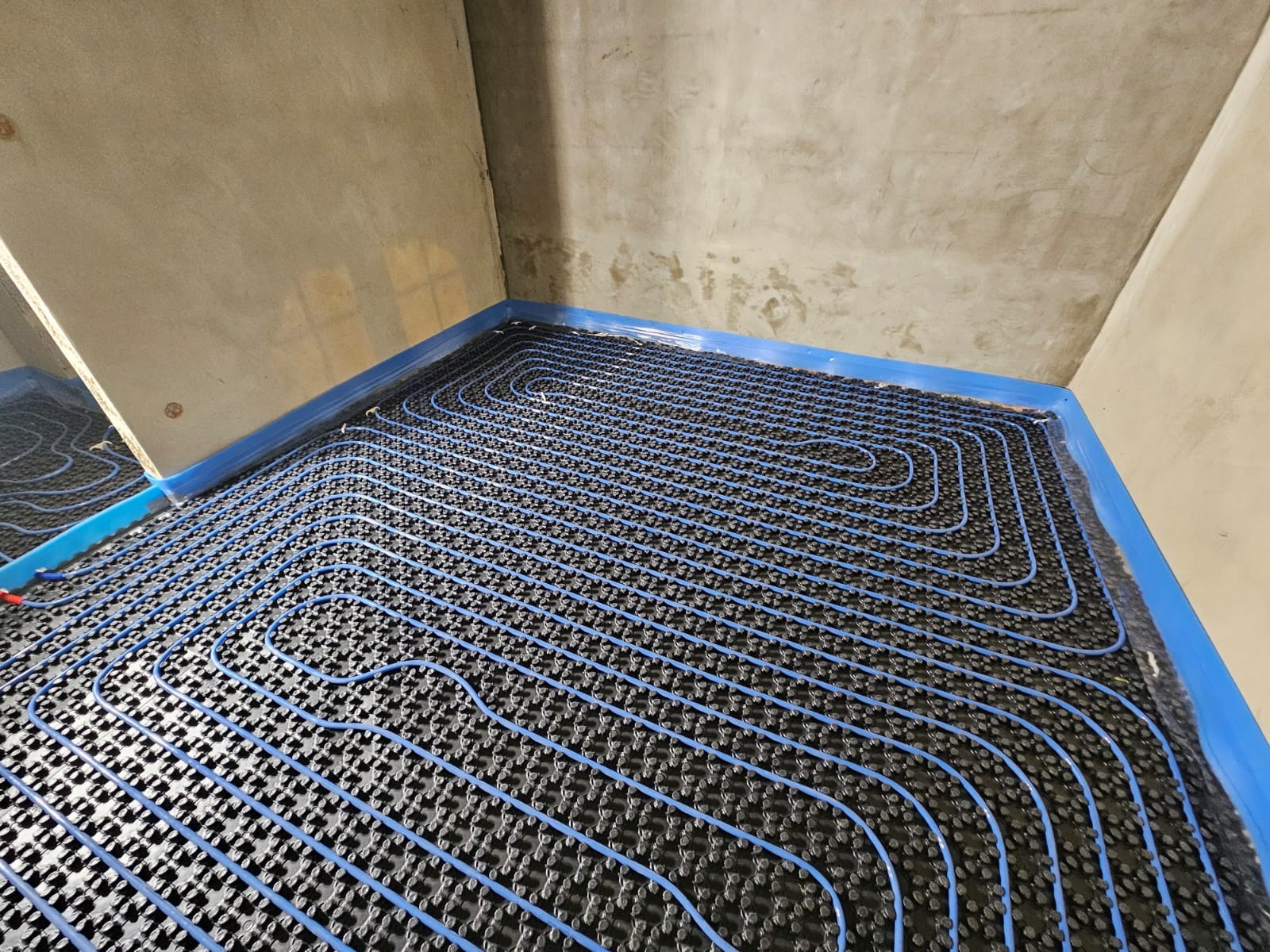 Niebieskie rury ogrzewania podłogowego ułożone na czarnej macie izolacyjnej, widok z góry na surową posadzkę w rogu pomieszczenia.