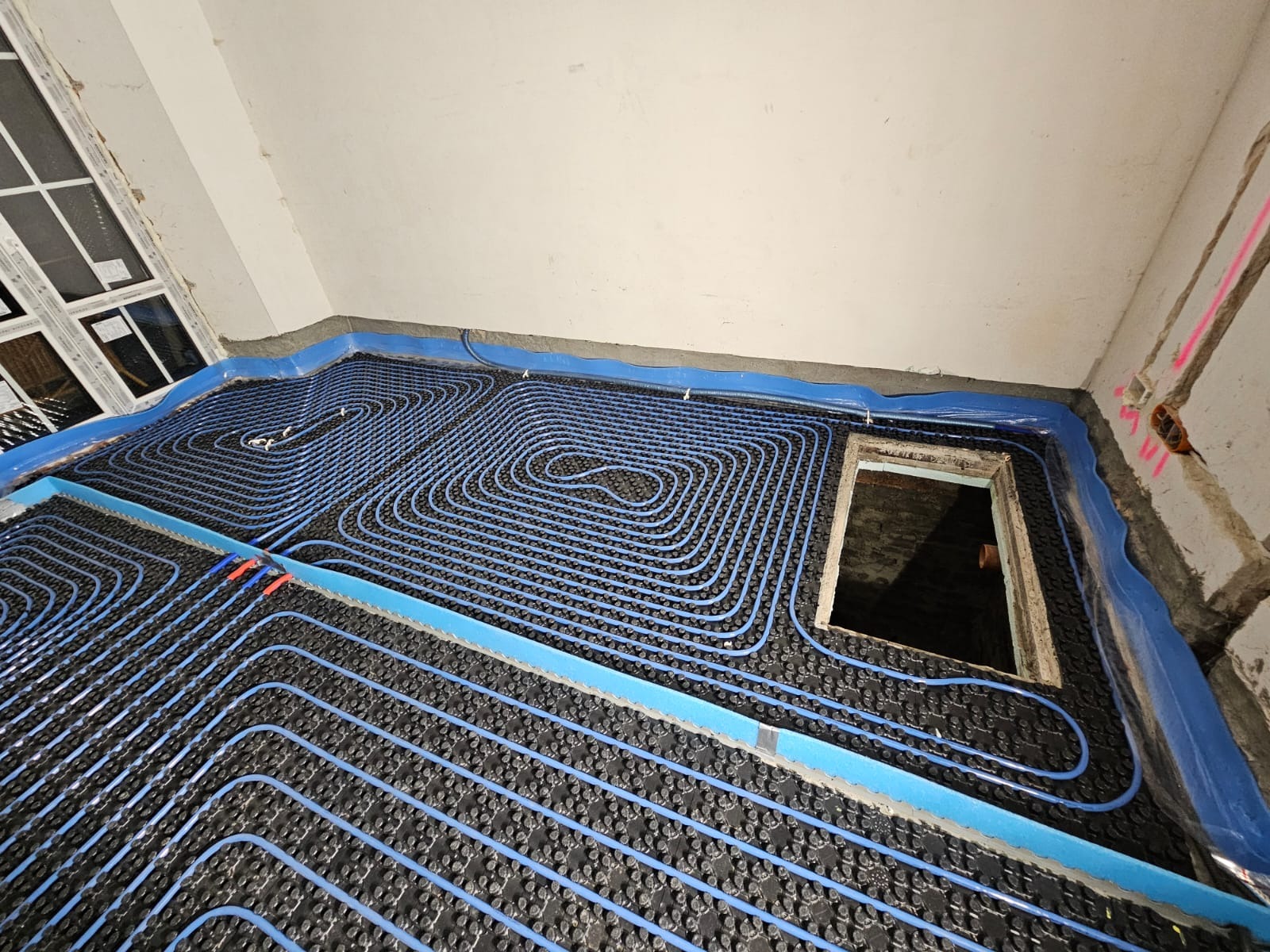 Niebieskie rurki ogrzewania podłogowego ułożone spiralnie na czarnej macie izolacyjnej w surowym pomieszczeniu z oknem i otworem w podłodze.