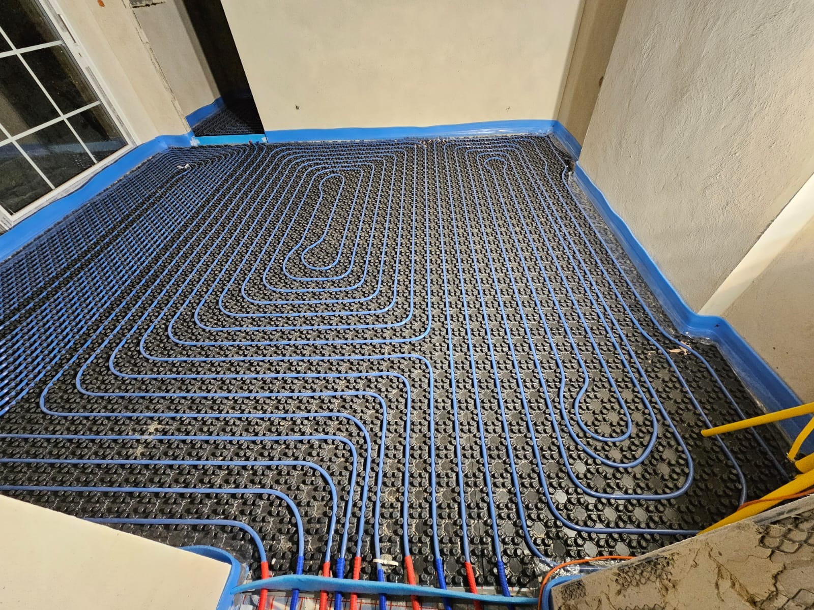 Niebieskie rury ogrzewania podłogowego ułożone spiralnie na czarnej macie izolacyjnej, zabezpieczone niebieską folią przy ścianach w surowym wnętrzu.