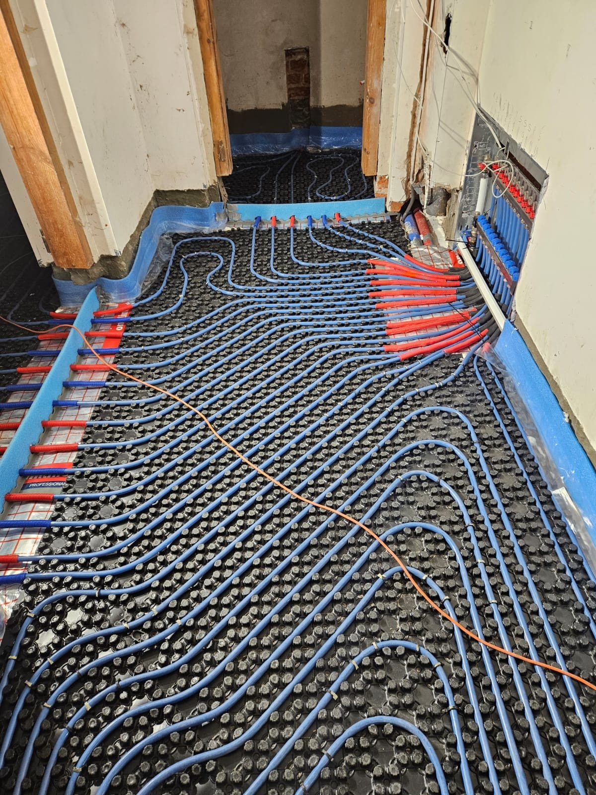 Niebieskie rury ogrzewania podłogowego ułożone na czarnej macie z wypustkami, widoczne czerwone końcówki rur i rozdzielacz, surowe ściany w tle.
