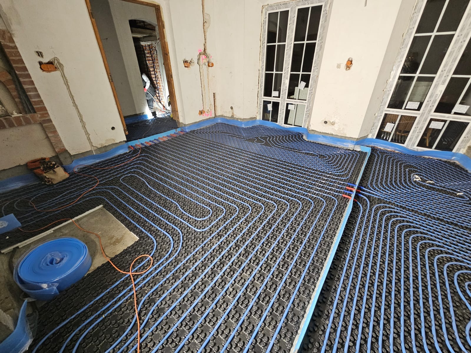 Niedokończona instalacja ogrzewania podłogowego: niebieskie rurki na czarnej macie, widoczne okna i przygotowane ściany do wykończenia.