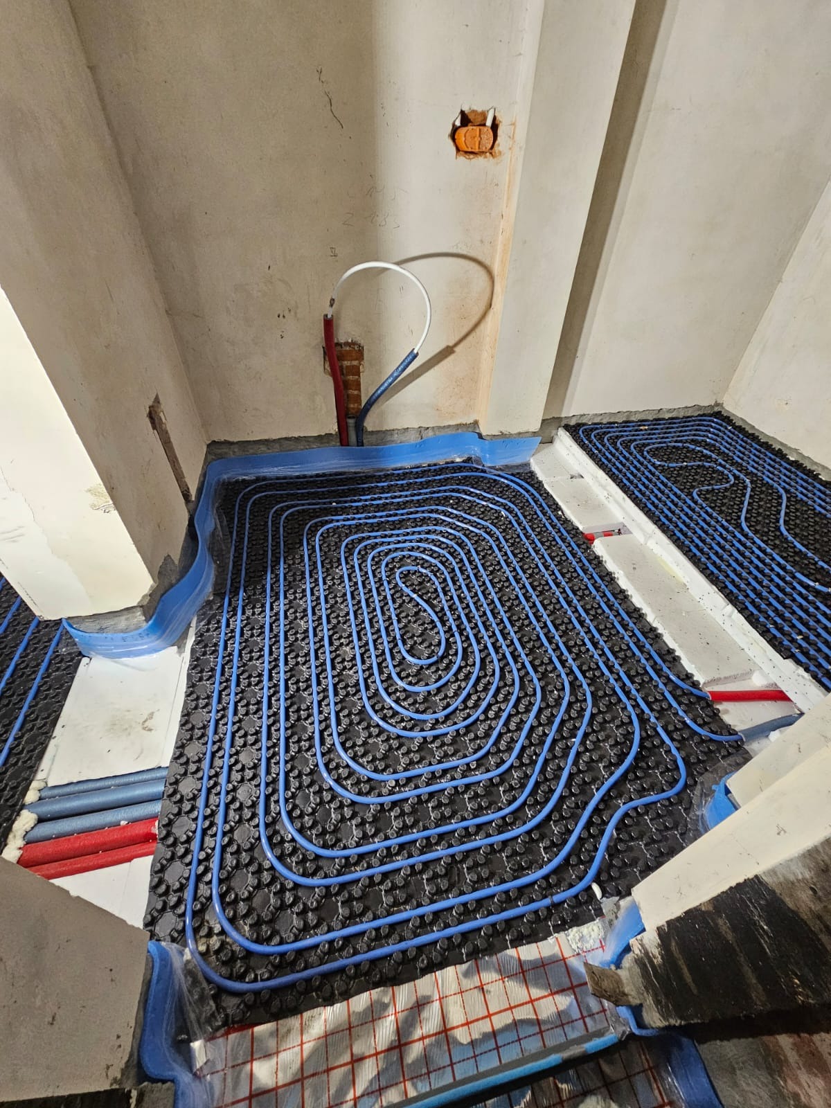 Niedokończona instalacja ogrzewania podłogowego z niebieskimi rurkami ułożonymi na czarnej macie izolacyjnej, widoczne fragmenty folii aluminiowej i rur doprowadzających, surowy stan ścian i podłogi.