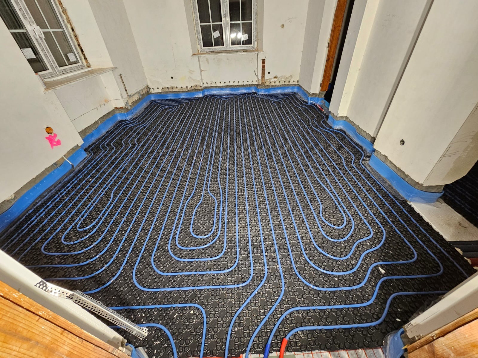 Niebieskie rurki ogrzewania podłogowego ułożone na czarnej macie izolacyjnej w pokoju z oknami i przygotowanymi ścianami do wykończenia.