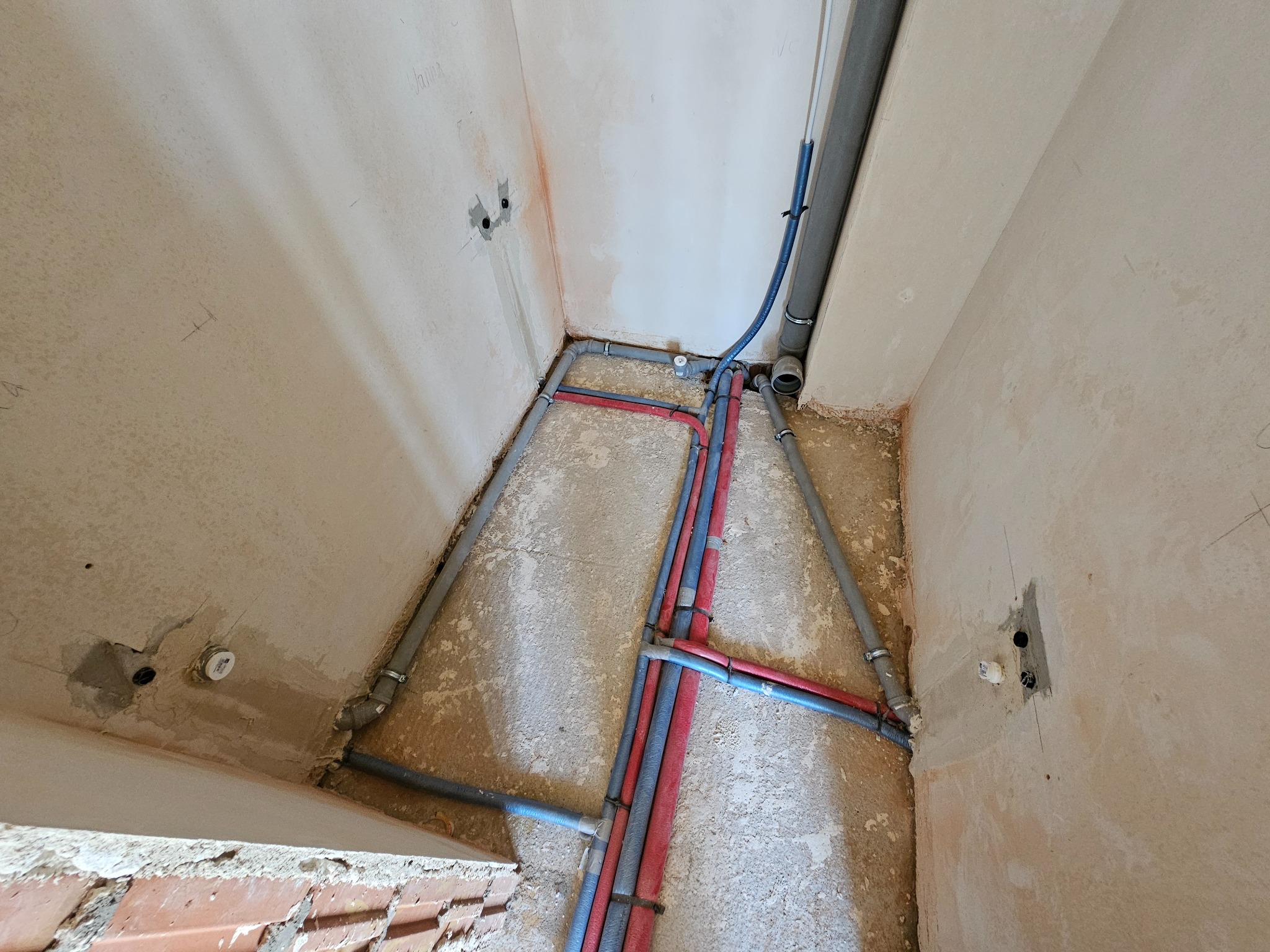 Instalacja wodna: rury w kolorze szarym, niebieskim i czerwonym ułożone w bruzdach w ścianie, widoczne uchwyty mocujące, ściany w surowym stanie.