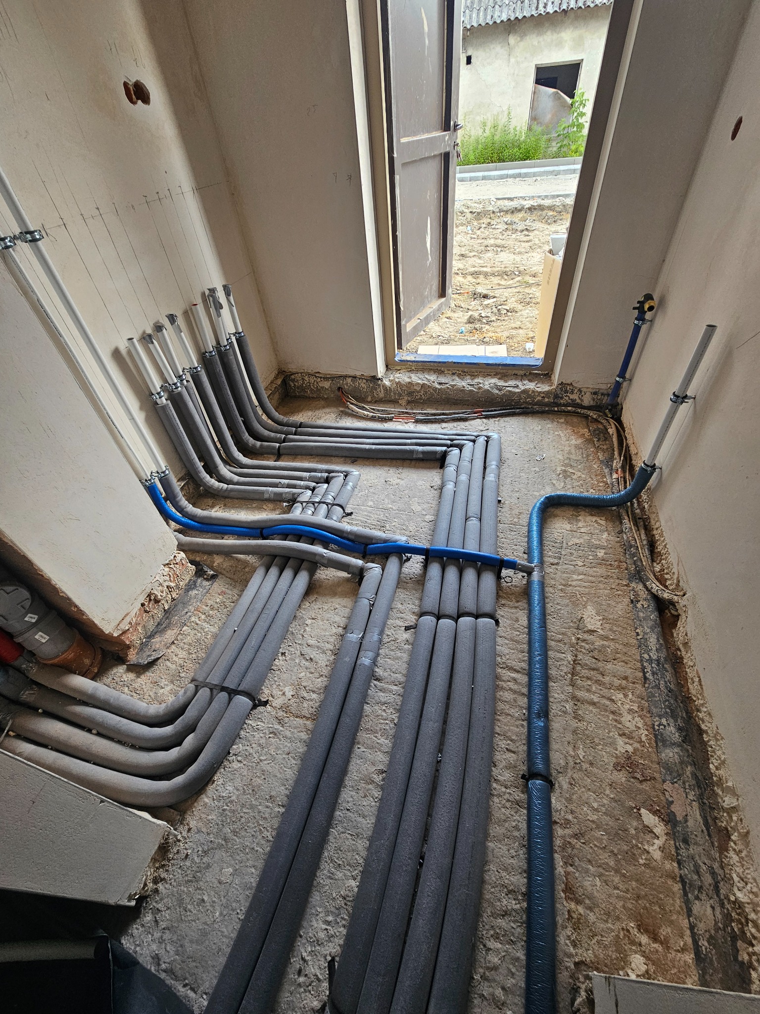 Instalacja rur w izolacji, ułożonych równolegle na podłożu w pomieszczeniu w trakcie remontu. Widoczne niebieskie rury oraz fragment otwartych drzwi.