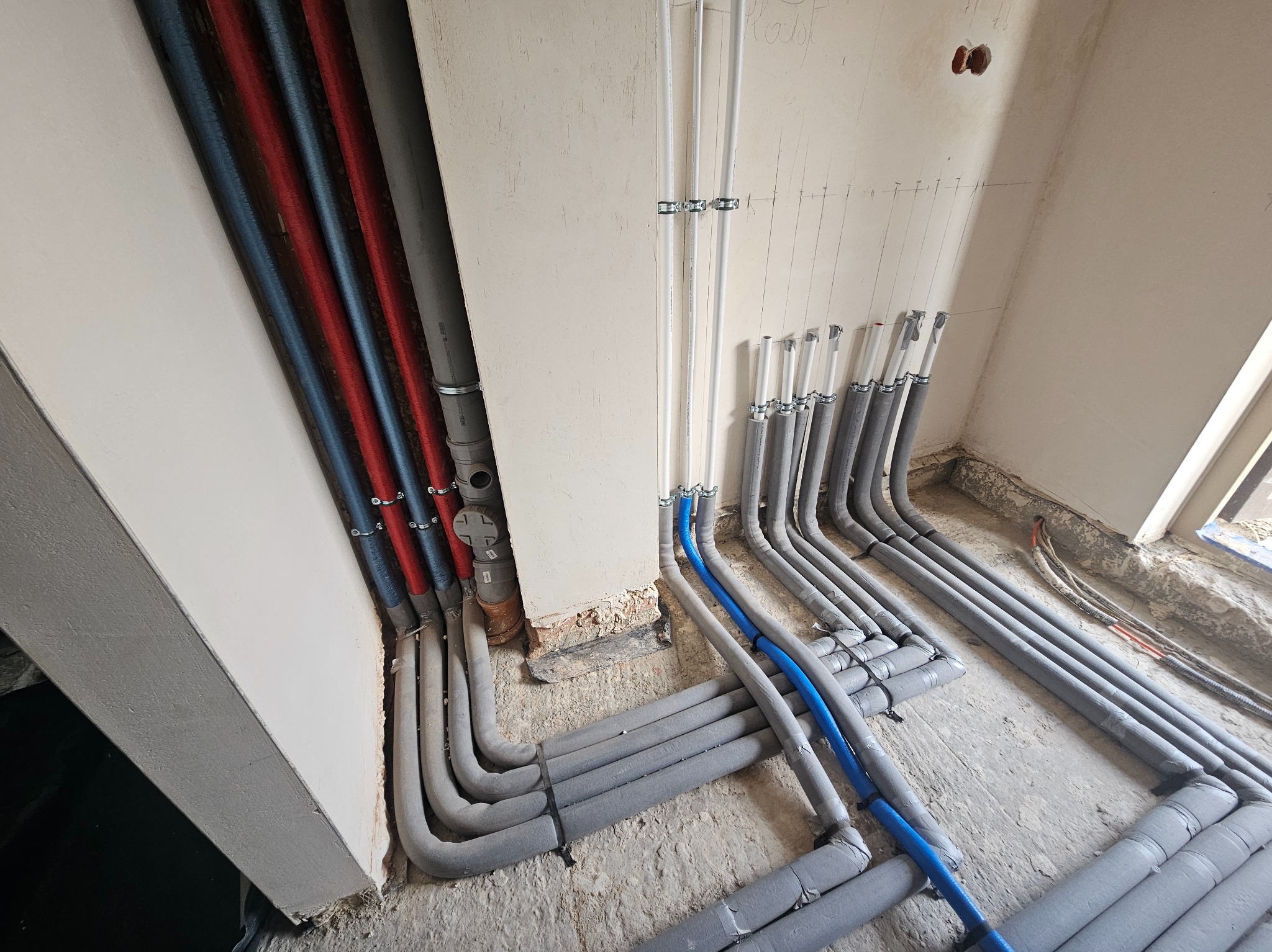 Szczegółowe ułożenie rur instalacyjnych w szarej izolacji, z niebieskimi i czerwonymi rurami w rogu pomieszczenia, widoczne mocowania na ścianie.