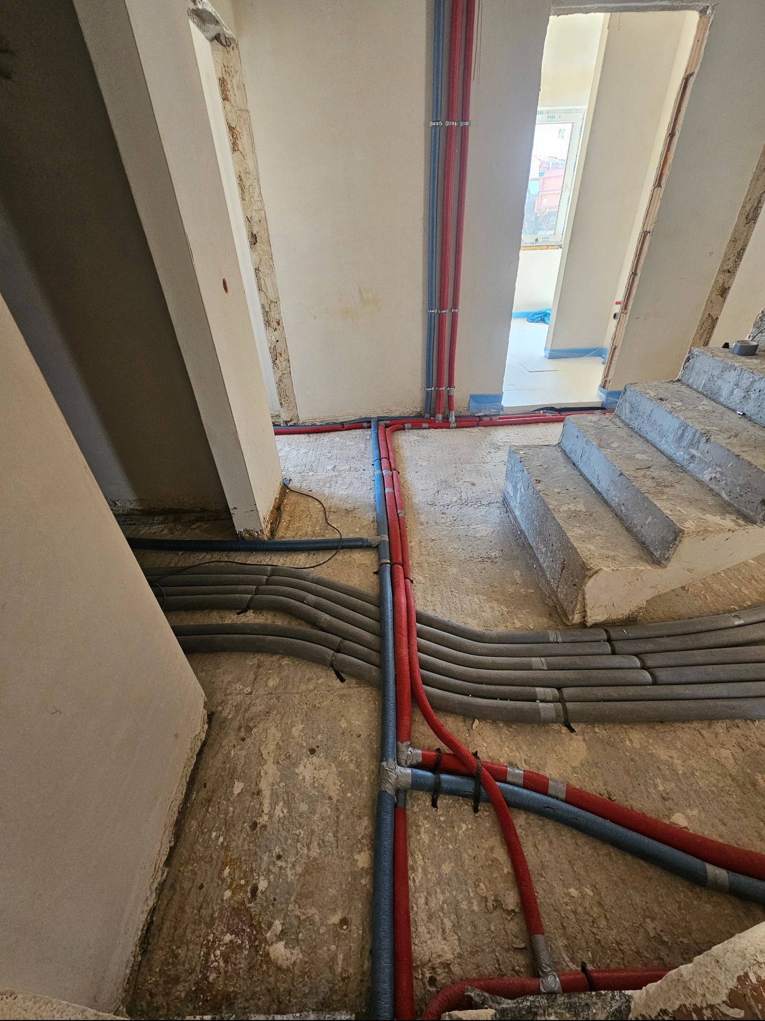 Instalacja grzewcza podłogowa w trakcie montażu: rury w izolacji termicznej ułożone w surowym stanie podłogi, widoczne fragmenty schodów i pionowe rury doprowadzające.