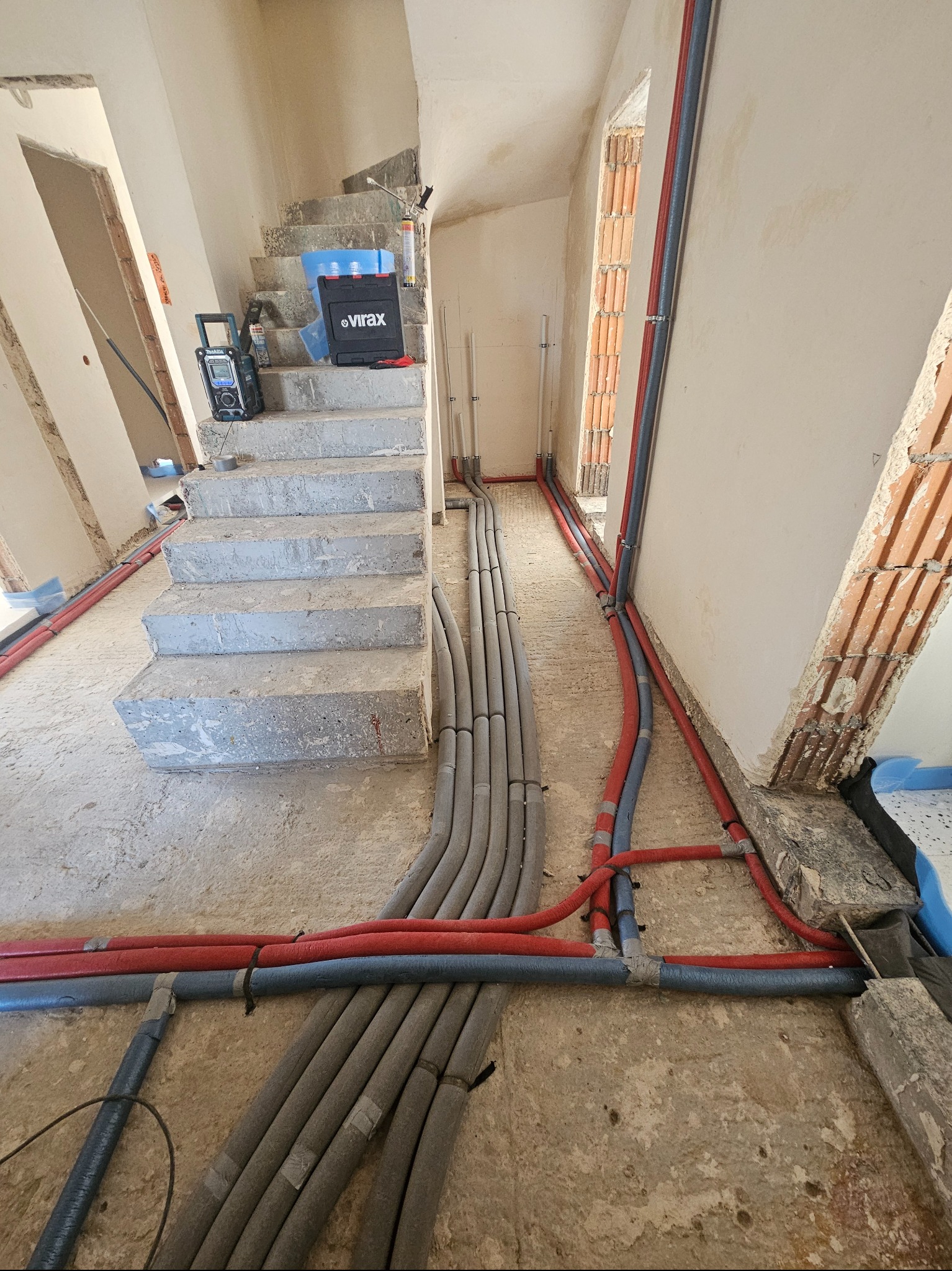 Instalacja rur grzewczych i wodnych w trakcie remontu domu w Warszawie. Widoczne szare i czerwone rury, narzędzia na schodach. Surowy stan.