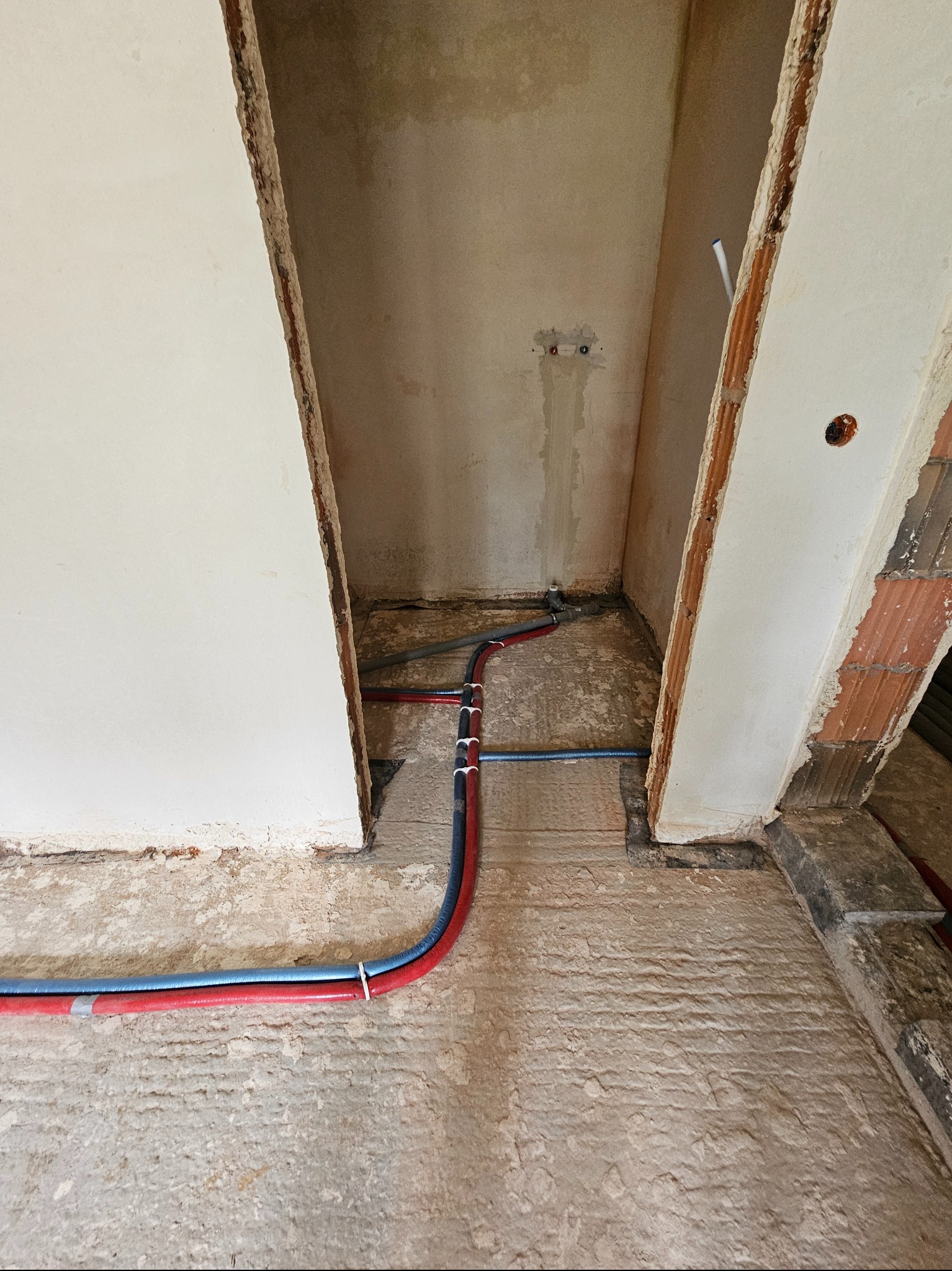 Rury instalacji wodnej (czerwona i niebieska) ułożone na surowym podłożu w pomieszczeniu w trakcie remontu. Widoczne ślady po kuciu w ścianie.