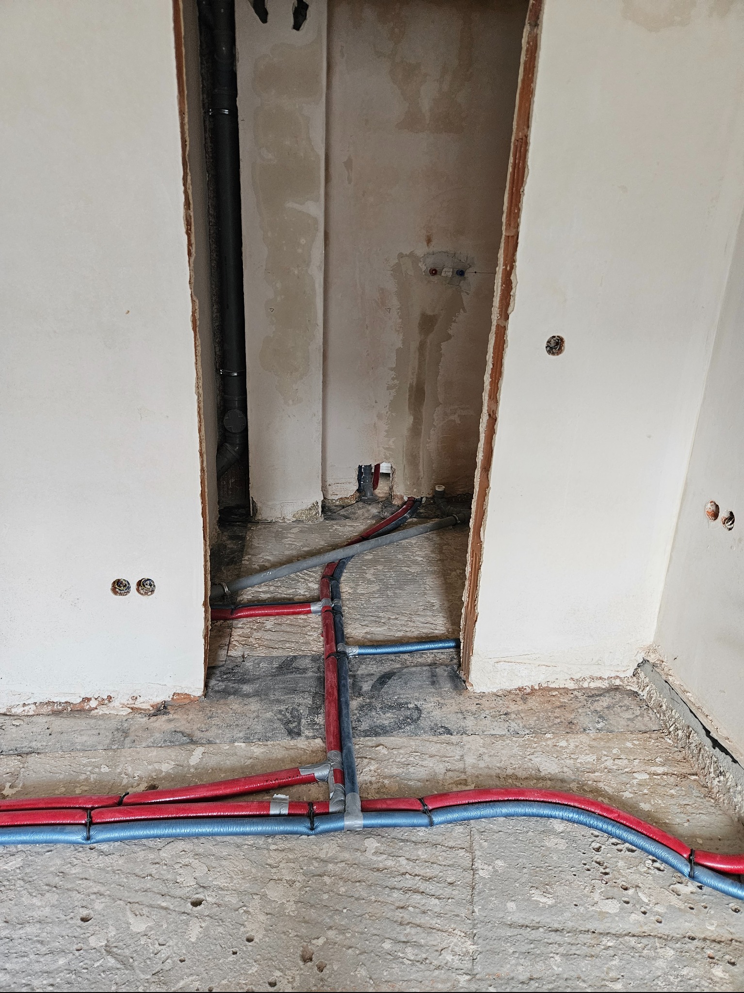 Instalacja rur w kolorze niebieskim i czerwonym na podłodze w surowym stanie. Widoczne otwory w ścianach i pionowa rura. Prace hydrauliczne w trakcie realizacji.