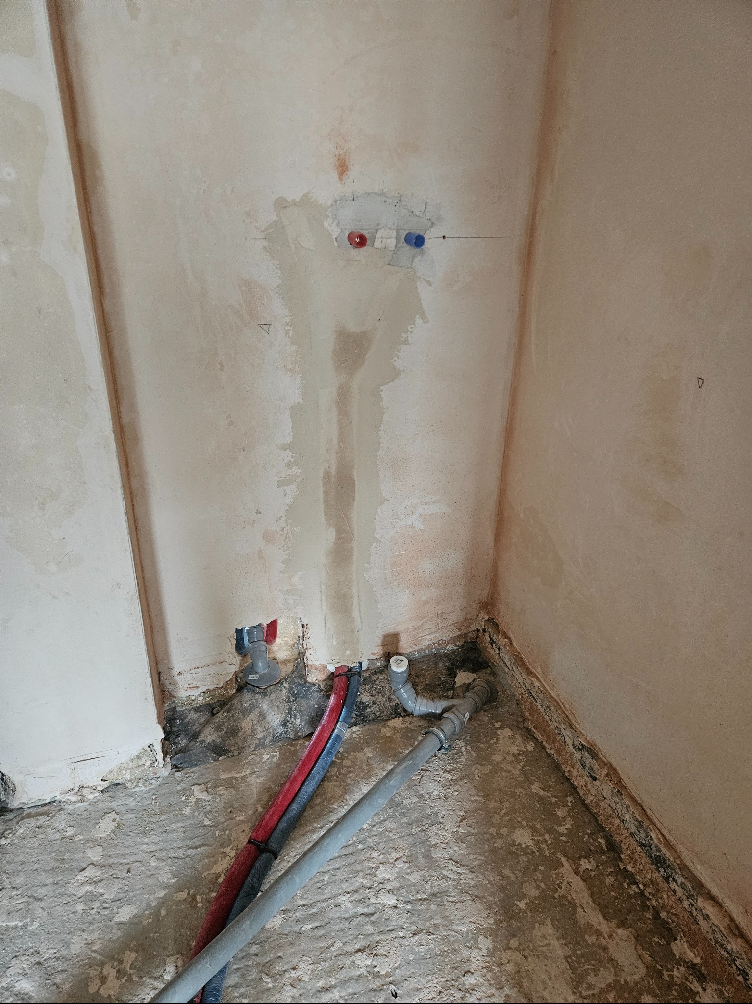 Fragment ściany z instalacją wodną w trakcie montażu. Widoczne rury, przyłącza i ślady po kuciu w tynku. Surowy stan wykończenia.