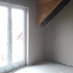 Świeżo otynkowane wnętrze poddasza z oknem PCV, widoczne elementy drewnianej konstrukcji dachu i przewody elektryczne przy podłodze.