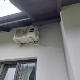 Biała jednostka zewnętrzna klimatyzacji Haier zamontowana na elewacji budynku, widoczne białe rury instalacyjne i fragment okna.