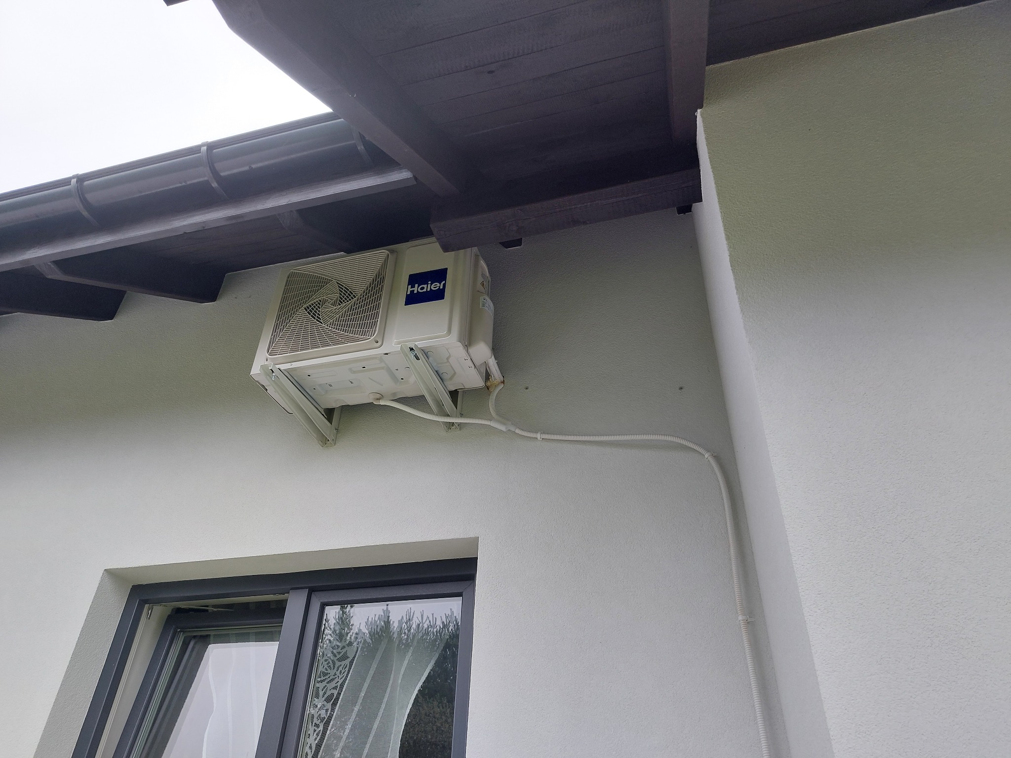 Biała jednostka zewnętrzna klimatyzacji Haier zamontowana na elewacji budynku, widoczne białe rury instalacyjne i fragment okna.