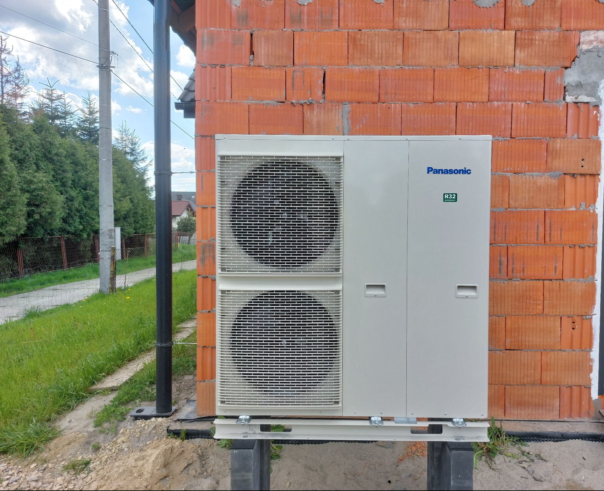 Zewnętrzna jednostka pompy ciepła Panasonic R32 zamontowana na betonowych blokach przy ścianie z czerwonej cegły dziurawki w trakcie budowy, widoczne niedociągnięcia w wykończeniu ściany,...