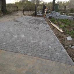 Układanie kostki brukowej na podjeździe, widoczny fragment ułożonej nawierzchni z szarej kostki, obok stos kostki, poziomica i brama wjazdowa.