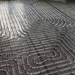 Impact Jakub Bauer - Układ rur ogrzewania podłogowego na folii izolacyjnej z czerwoną kratką, widok z góry w trakcie montażu.