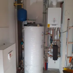 Kotłownia w trakcie montażu: biały zasobnik ciepłej wody owinięty folią, piec gazowy, rury miedziane i stalowe, manometry, drabina oparta o ścianę.